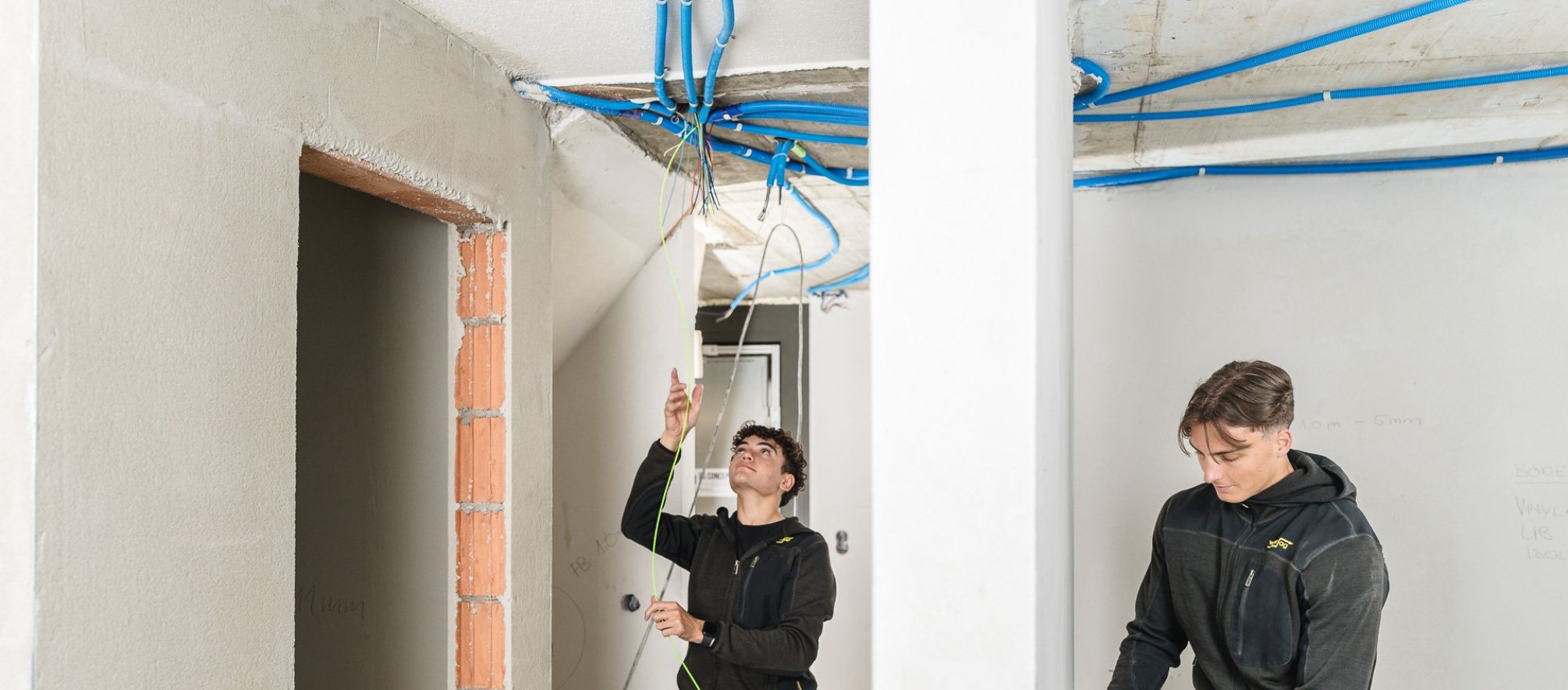 Zwei Elektroinstallateure verlegen und verbinden Kabel in einem Rohbau. Blaue Installationsrohre verlaufen an der Decke, w&auml;hrend die Arbeiten an der Elektroinstallation in den Innenr&auml;umen ausgef&uuml;hrt werden.